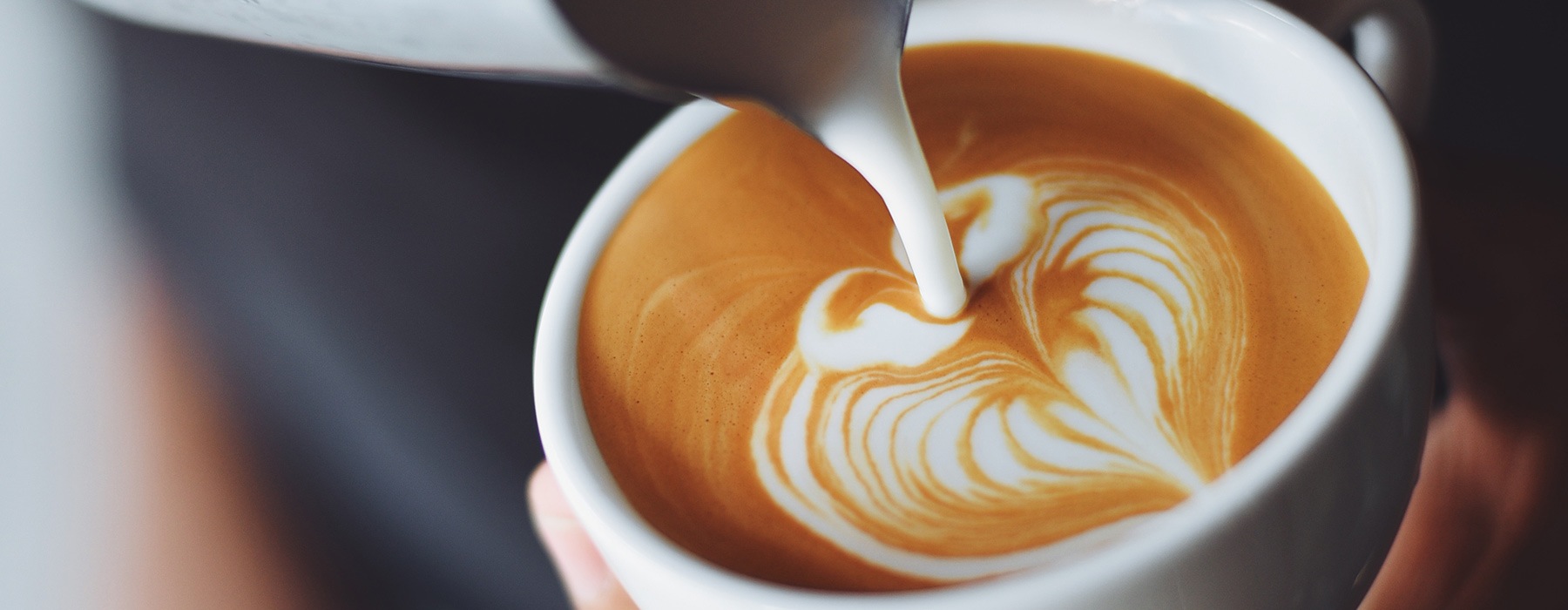 close-up image of cream being poured into latte art on the top of a hot coffee drink